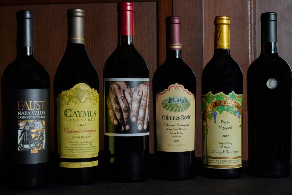 Six bottles of red wine are lined up side by side on a wooden surface, each with distinct labels, including brands like Faust, Caymus Vineyards, and Chimney Rock. The bottle on the right has no visible label.
