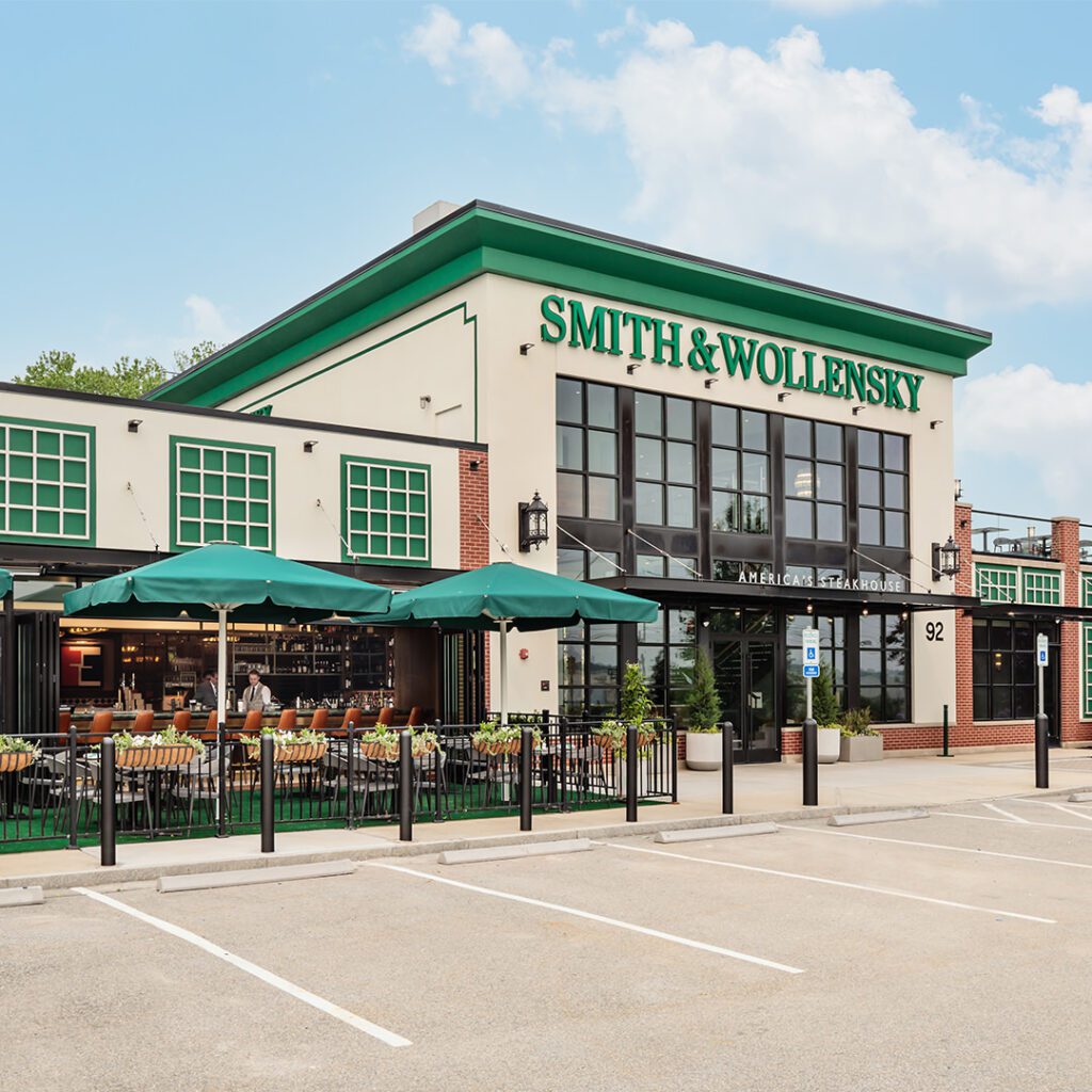 Smith & Wollensky steakhouse with green accents, large windows, and outdoor seating under green umbrellas. The restaurant name is prominently displayed above the entrance. Several empty parking spaces are in front.