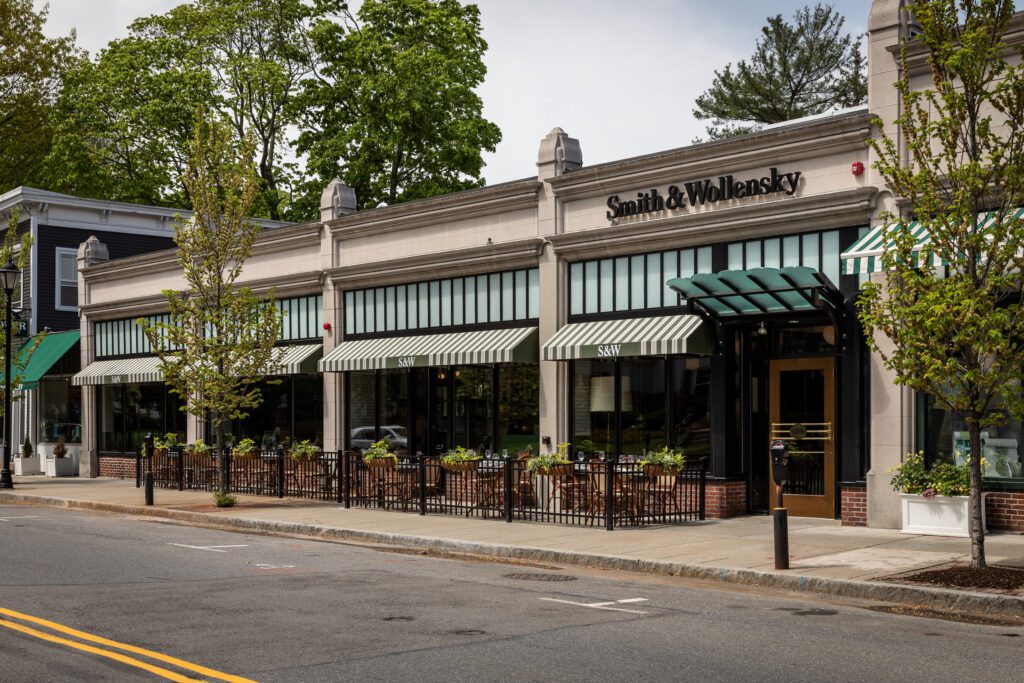 Street view of a Smith & Wollensky restaurant with striped green and white awnings, outdoor patio seating, potted plants, and trees, on a quiet road with no visible cars or people.