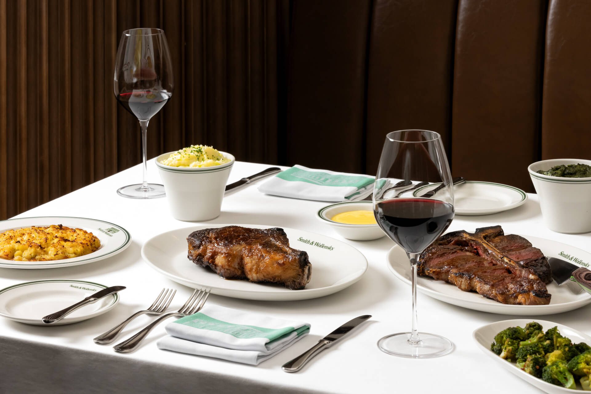 Restaurant table with plates of steak, broccoli, mac and cheese, and creamed spinach. Two glasses of red wine and a side of sauce are also present. White tablecloth and napkins complete the setting.