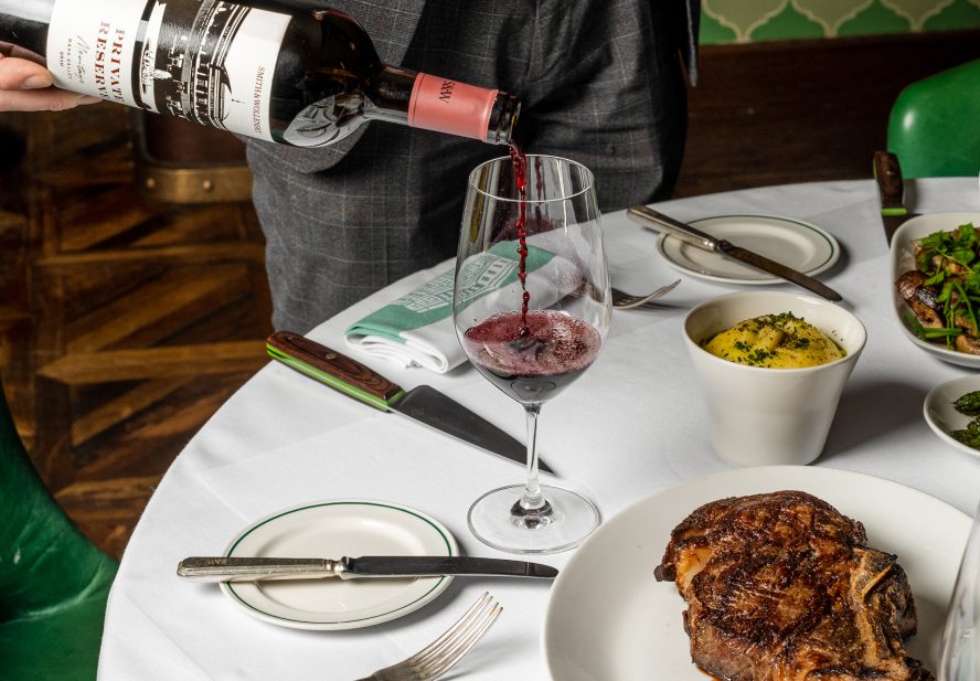 A person pours red wine into a glass on a white tablecloth set with a grilled steak, a bowl of mashed potatoes, leafy greens, and utensils.