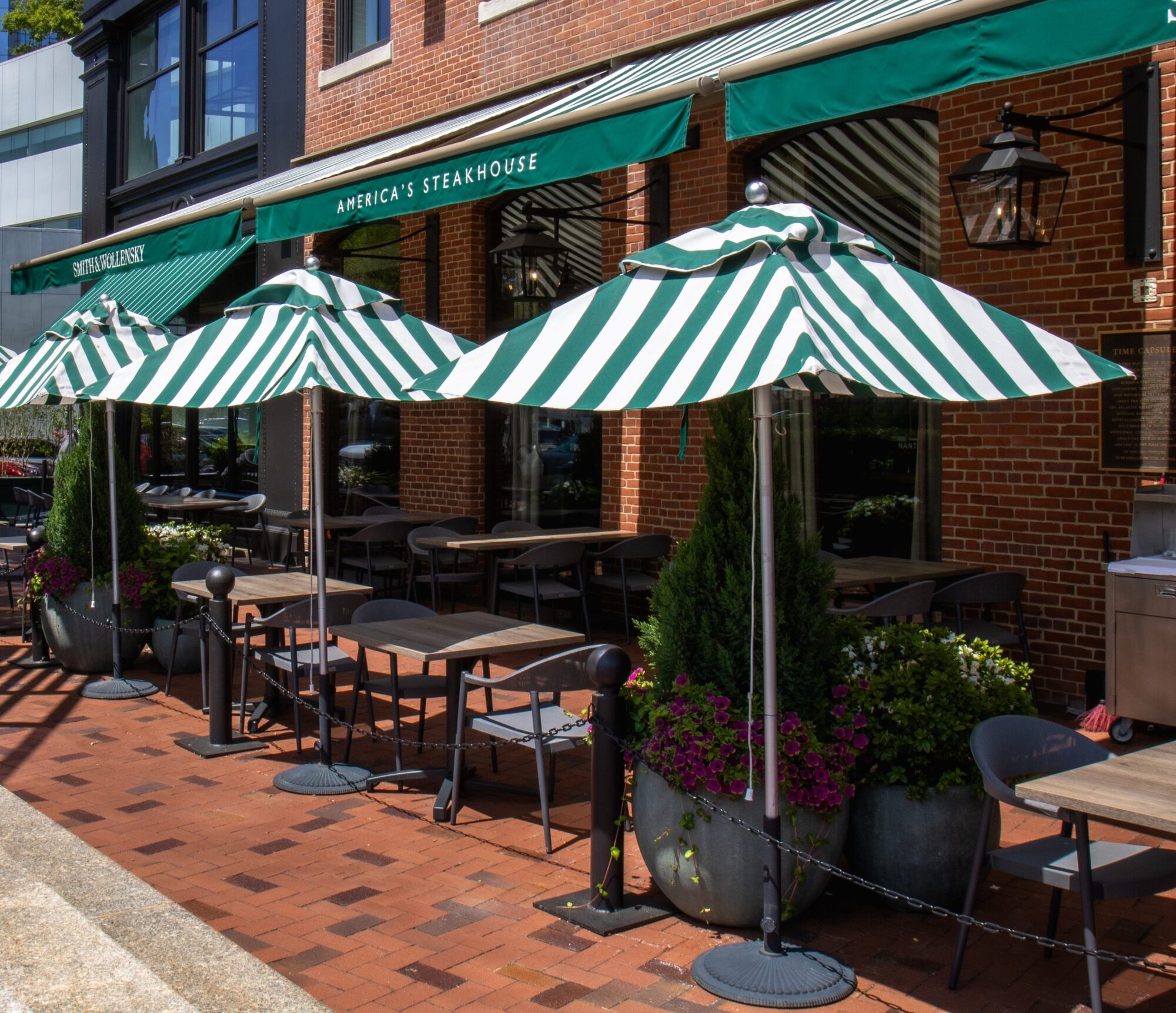 Outdoor seating area of a steakhouse with green and white striped umbrellas, potted plants, and empty tables and chairs on a brick sidewalk in front of a red brick building.
