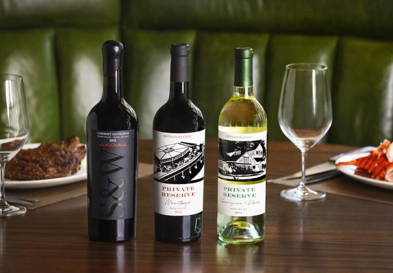 Three bottles of wine are arranged on a wooden table, flanked by empty wine glasses. Plates of steak and lobster are partially visible in the background, with green upholstered seating behind.