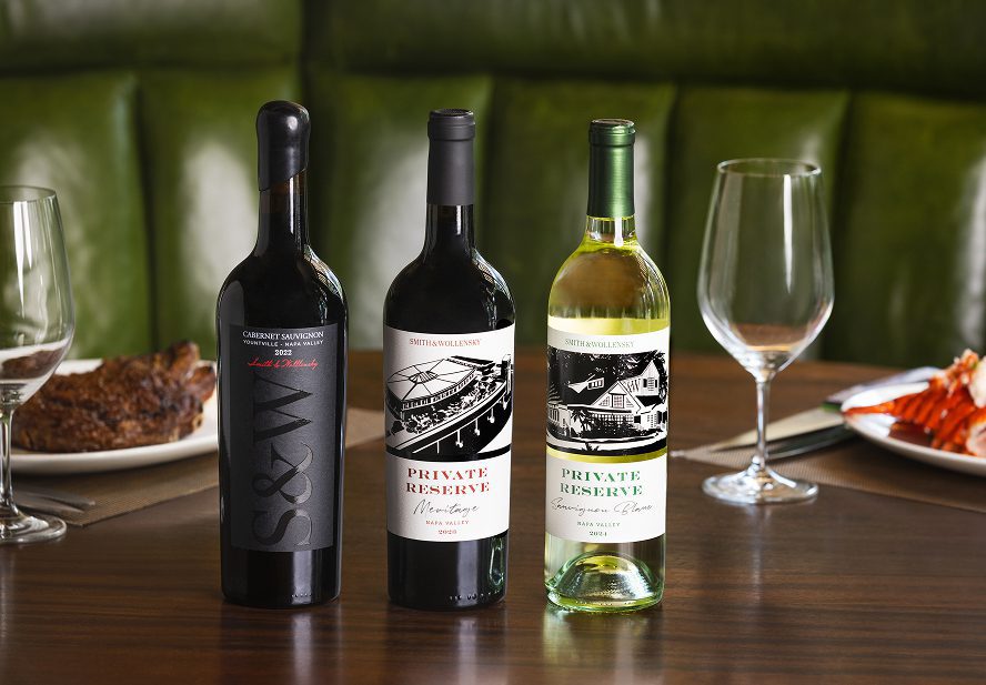 Three bottles of wine are arranged on a wooden table, flanked by empty wine glasses. Plates of steak and lobster are partially visible in the background, with green upholstered seating behind.