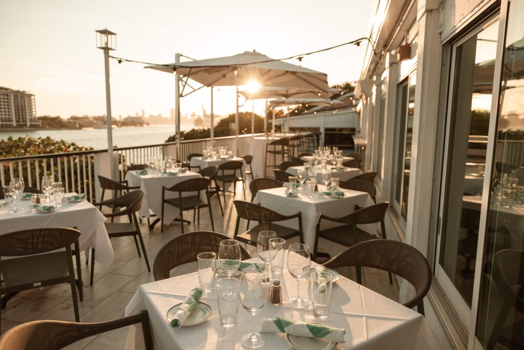 Outdoor restaurant patio with white tablecloths and neatly set tables at sunset, overlooking the water and city skyline. Sunlight shines through umbrellas, creating a warm and inviting atmosphere.