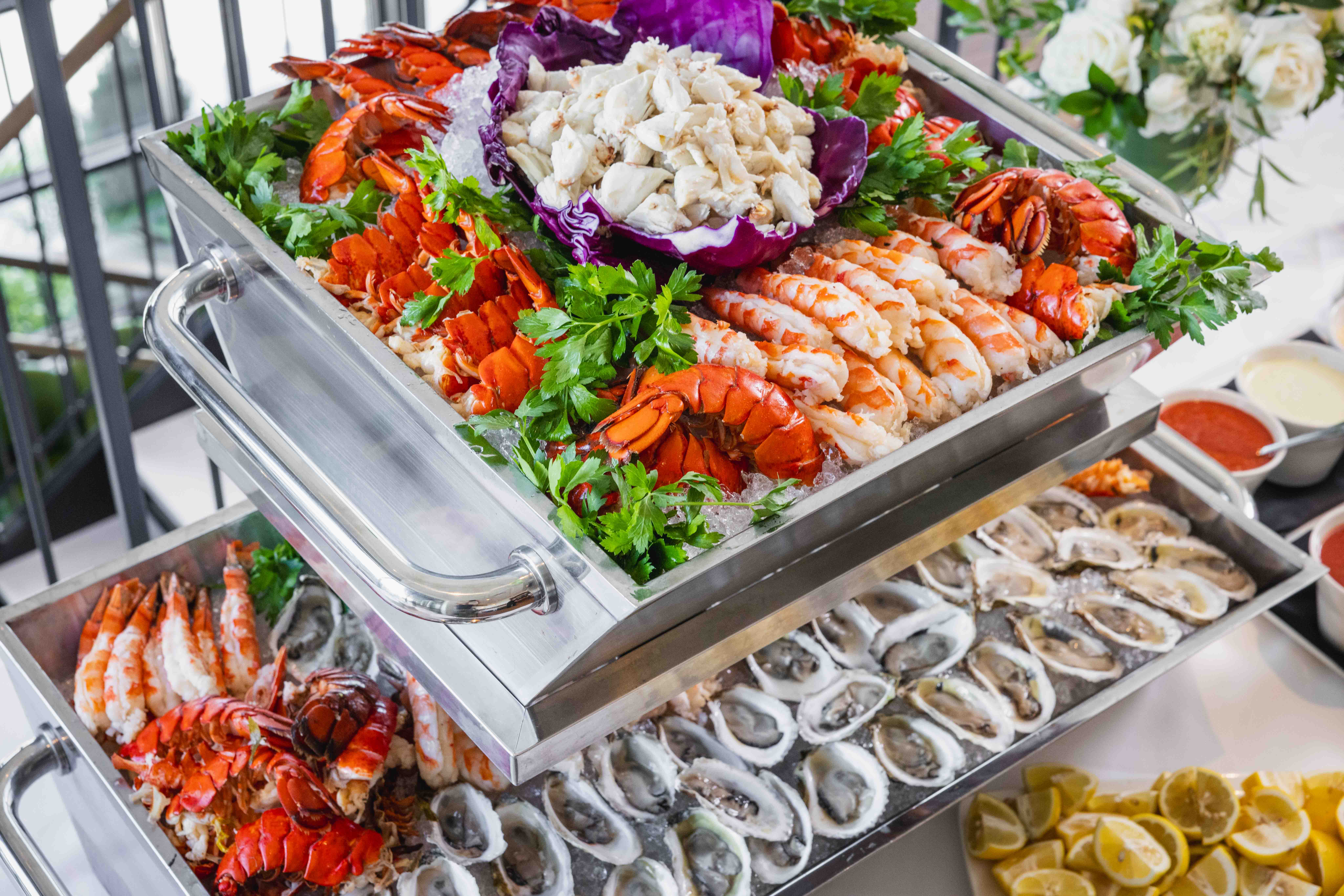 A buffet display of seafood including lobster tails, shrimp, and crab meat on ice, garnished with parsley, and a lower tray filled with oysters on the half shell. Lemon wedges and sauces are on the side.