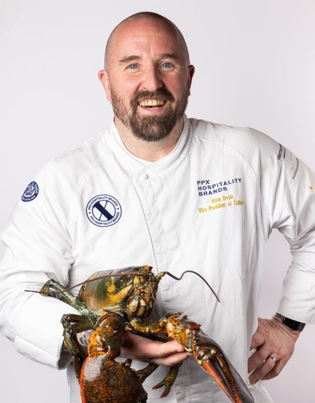 A smiling chef in a white uniform holds a large lobster with both hands. The chef has a beard and is standing against a plain white background.