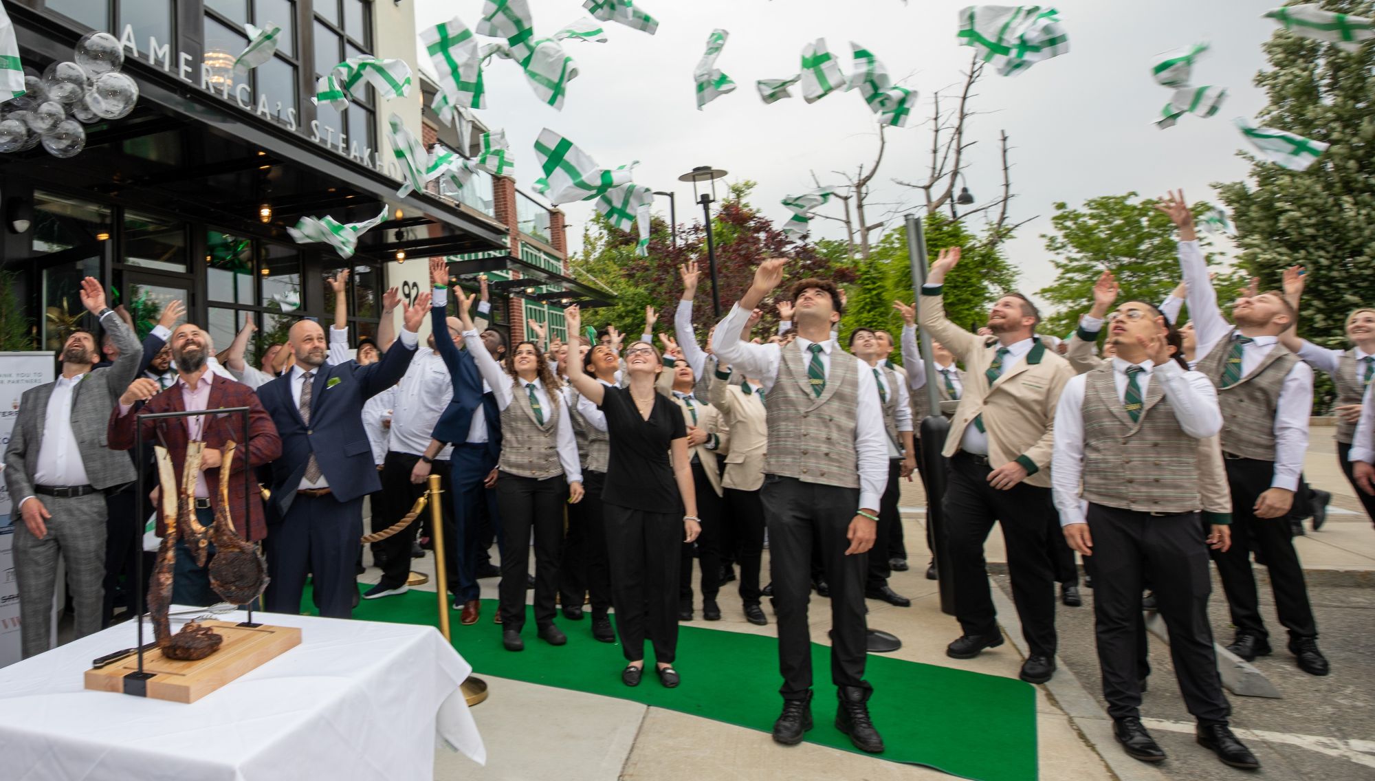 A group of people in formal attire stand outside a building, tossing green-and-white objects into the air and celebrating. Some smile and look up, while a table with a ham leg and knife is visible in the foreground.