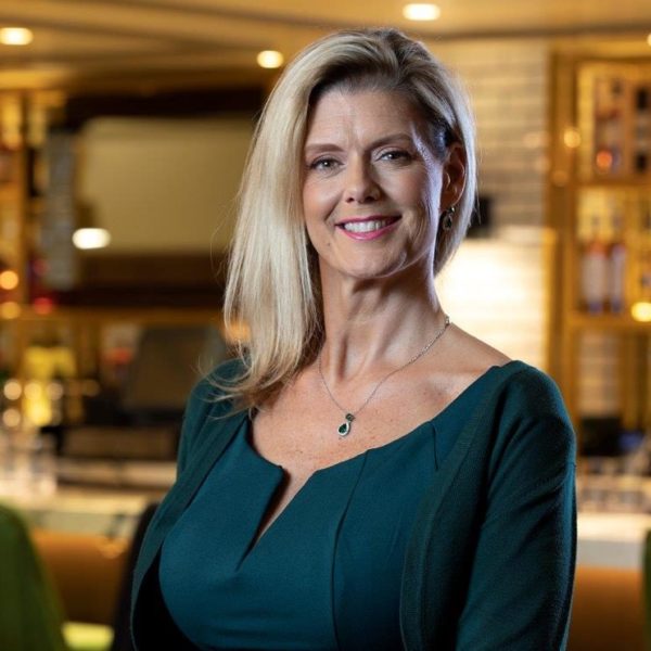 A woman with long blonde hair, wearing a green dress and a pendant necklace, smiles while standing in a warmly lit indoor setting with blurred shelves and bottles in the background.