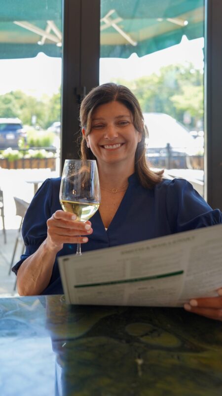 A woman sitting at a restaurant table, smiling, holding a glass of white wine in one hand and a menu in the other, with sunlight streaming through large windows behind her.