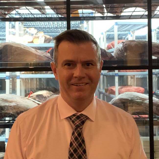 A man in a pink shirt and plaid tie smiles in front of shelves filled with large cuts of meat in a refrigerated storage area.