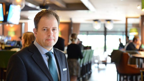 A man in a suit and tie stands in a modern, softly lit restaurant or café. The background shows people sitting at tables and a bar, with a relaxed and professional atmosphere.