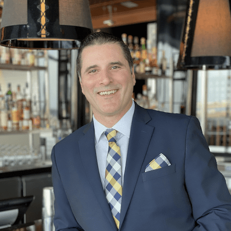 A man in a dark suit with a patterned tie and pocket square smiles while standing in a modern bar with bottles and glasses on shelves in the background.