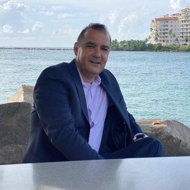 A man in a dark suit and light pink shirt sits on rocks by the water, smiling. In the background, there are waterfront buildings, trees, and a partly cloudy sky.