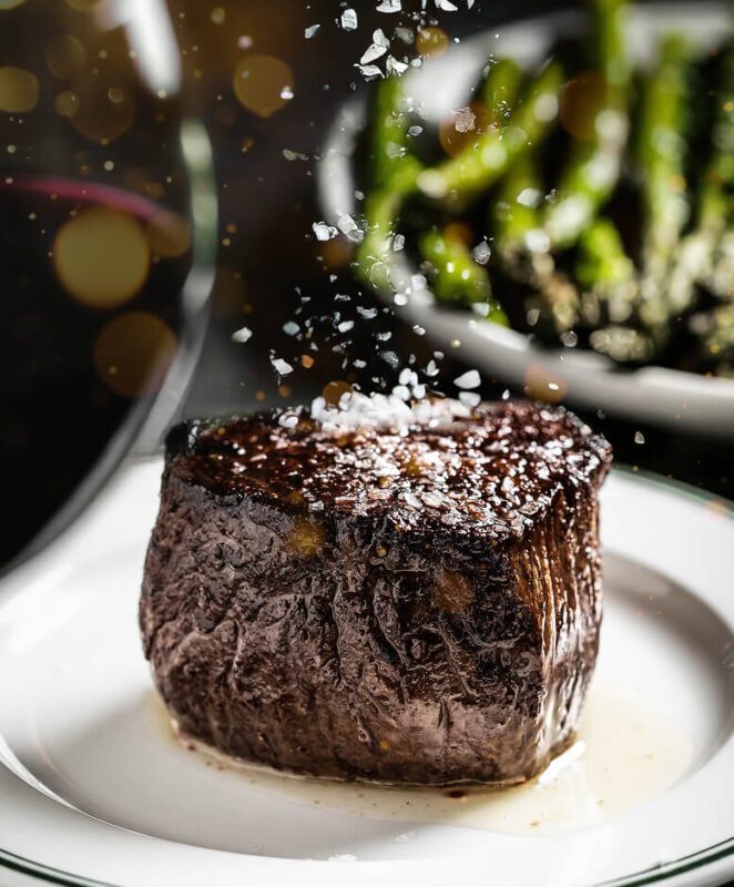 A perfectly cooked filet mignon steak on a white plate, sprinkled with coarse sea salt, with a blurred background featuring green asparagus and a glass of red wine.