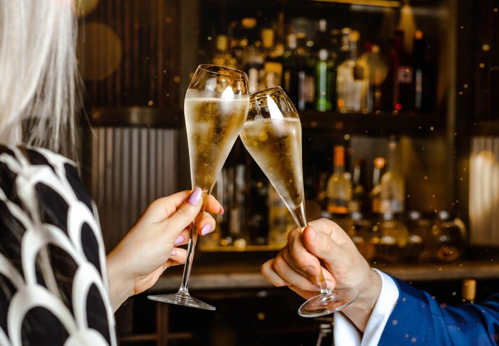 Two people clink champagne glasses in a toast at a bar, with bottles visible in the background. One person wears a patterned outfit, the other a blue suit.