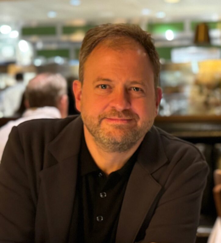 A man with short light brown hair and a beard, wearing a black shirt and dark blazer, sits at a table in a warmly lit restaurant, smiling softly at the camera. The background shows blurred diners and lights.