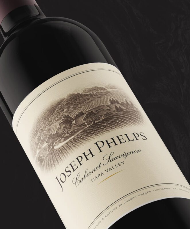 A close-up of a wine bottle labeled “Joseph Phelps Cabernet Sauvignon Napa Valley,” featuring an illustration of vineyards and hills on the cream-colored label against a dark background.