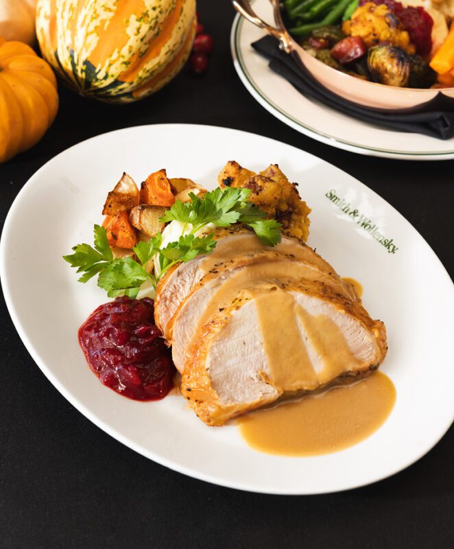 A plate with sliced turkey topped with gravy, cranberry sauce, stuffing, and roasted vegetables, garnished with parsley. In the background are pumpkins and a dish of more vegetables.