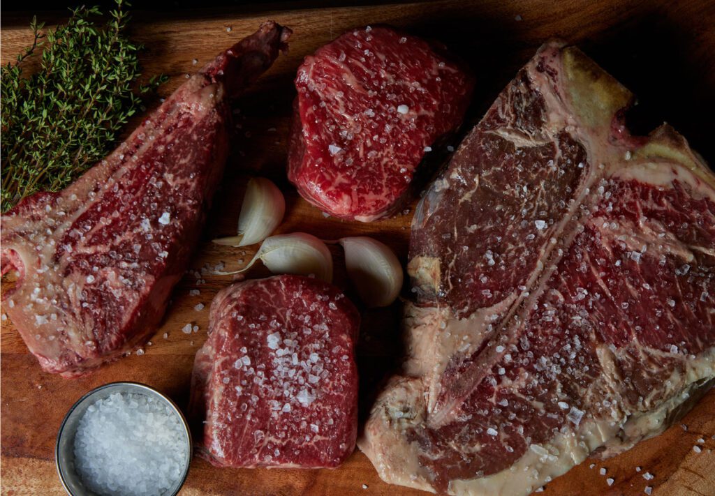 Various cuts of raw steak sprinkled with coarse salt are arranged on a wooden board, along with sprigs of thyme, garlic cloves, and a small bowl of salt.