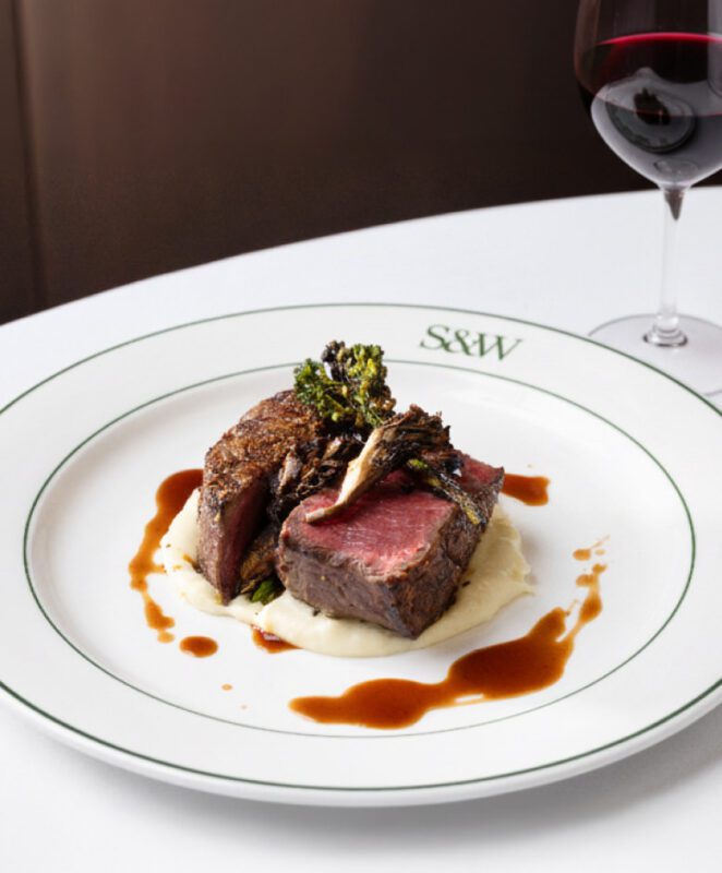 A plate with two pieces of medium-rare steak on mashed potatoes, garnished with grilled vegetables and brown sauce, sits on a white tablecloth beside a glass of red wine.
