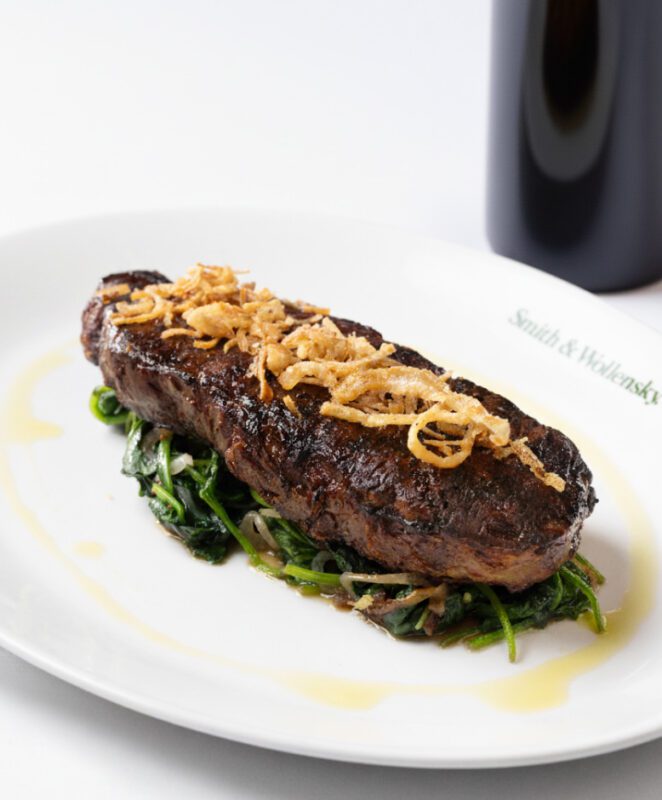 A grilled steak topped with crispy fried onions sits on sautéed spinach, served on a white plate with a light drizzle of sauce. The plate is labeled 