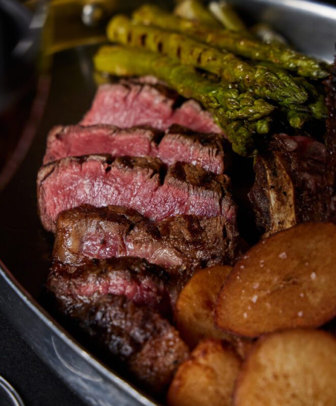 A close-up of sliced, medium-rare steak served with grilled asparagus and roasted potato slices on a metal platter.