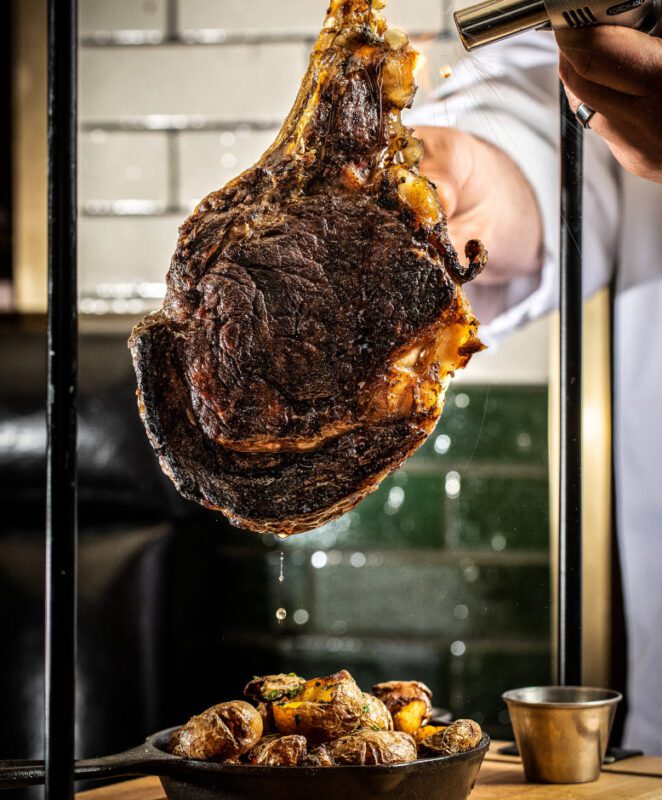 A large, juicy tomahawk steak is held above a skillet of roasted potatoes while a hand uses a blowtorch to sear the meat. Juices drip down onto the potatoes, and a small metal cup sits nearby.