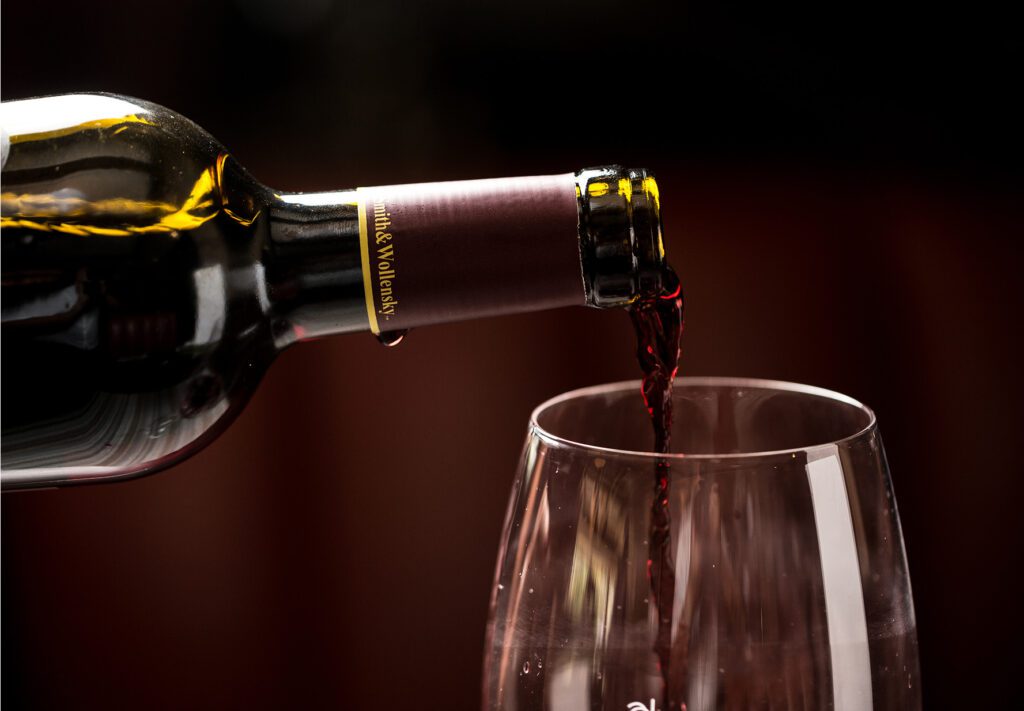 A bottle of red wine is being poured into a clear wine glass against a dark, blurred background.
