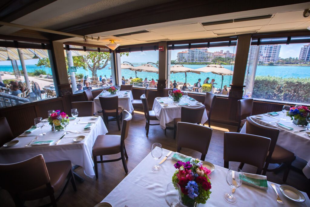 Elegant restaurant dining area with tables set for service, floral centerpieces, and large windows offering a view of an outdoor patio, blue water, and distant buildings under a sunny sky.