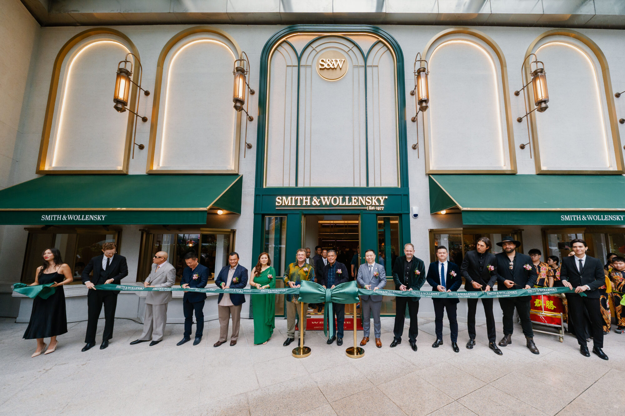 A group of people in formal attire stand in front of Smith & Wollensky restaurant, holding a large green ribbon for a grand opening ceremony; green awnings and gold accents decorate the entrance.