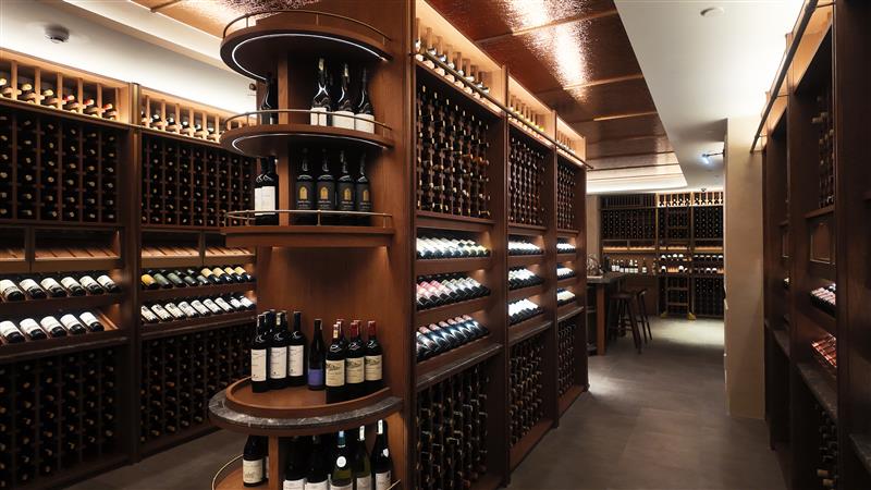 A well-lit wine cellar with wooden shelves and racks filled with bottles of wine, featuring a central round display and a seating area with stools in the background.