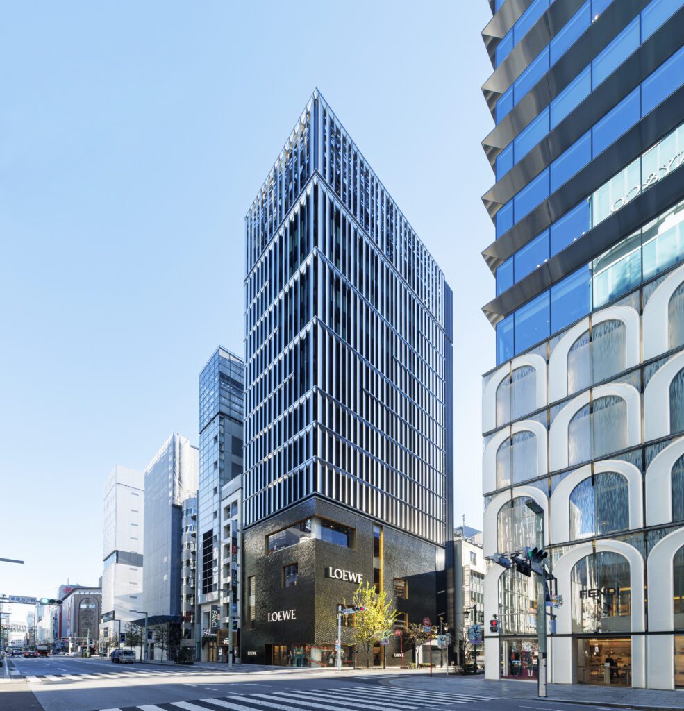 A modern city street with tall buildings, including a black multi-story Loewe store with geometric patterns on its facade, under a clear blue sky. The street is empty and sunlight illuminates the scene.
