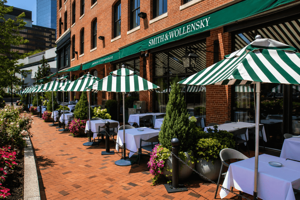 Outdoor dining area at Smith & Wollensky restaurant with green-and-white striped umbrellas, white tablecloth-covered tables, potted plants, and brick walkway on a sunny day. Brick building facade and city buildings in background.