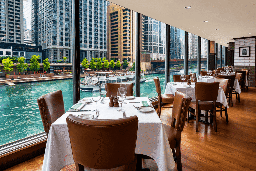 Elegant restaurant with brown leather chairs and white tablecloths, set beside large windows overlooking a river and tall city buildings on a sunny day.