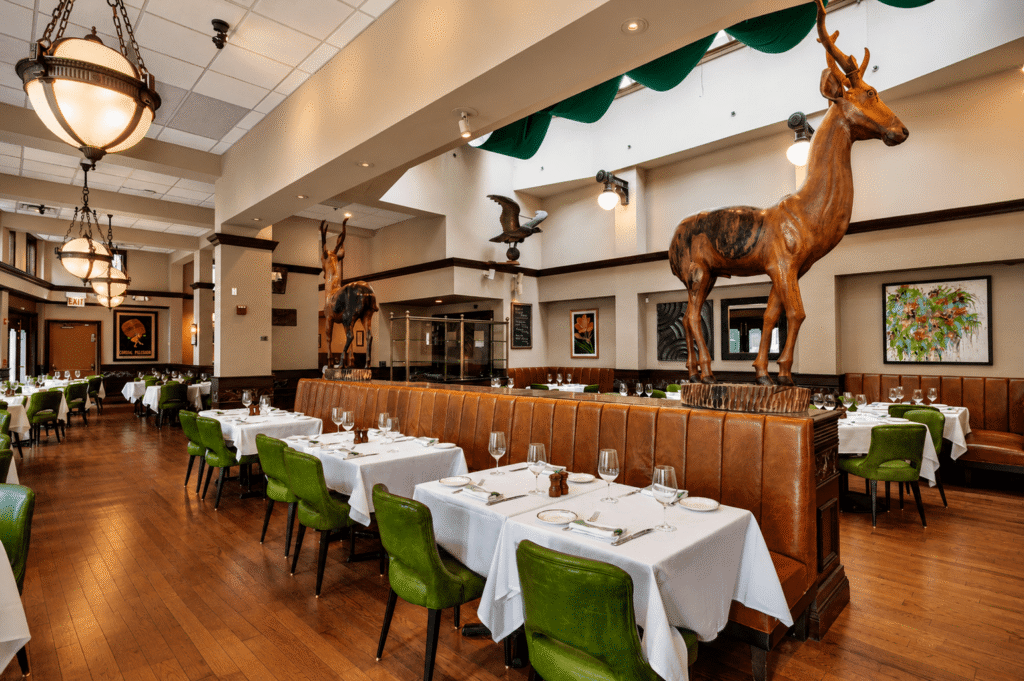 Upscale restaurant interior with green chairs, white tablecloths, and large animal sculptures, including deer, displayed on booths. Wooden floors, art on the walls, and modern lighting create a stylish yet inviting atmosphere.