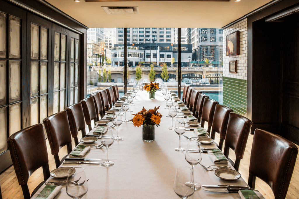 Elegant dining room with a long table set for a formal meal, featuring brown leather chairs, white tablecloth, flower centerpieces, glassware, and a cityscape view through large windows at the far end.