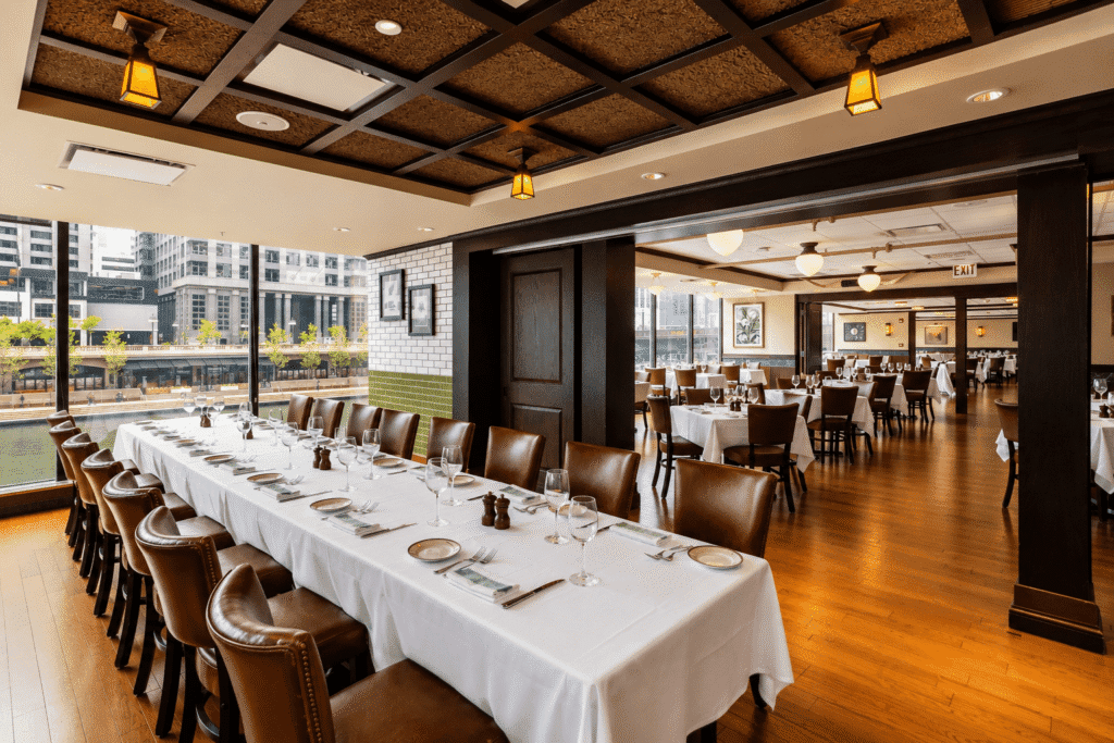 Elegant restaurant interior with large windows, wooden floors, brown leather chairs, and tables set with white tablecloths and glassware. The space is bright, spacious, and overlooks a cityscape with modern buildings.