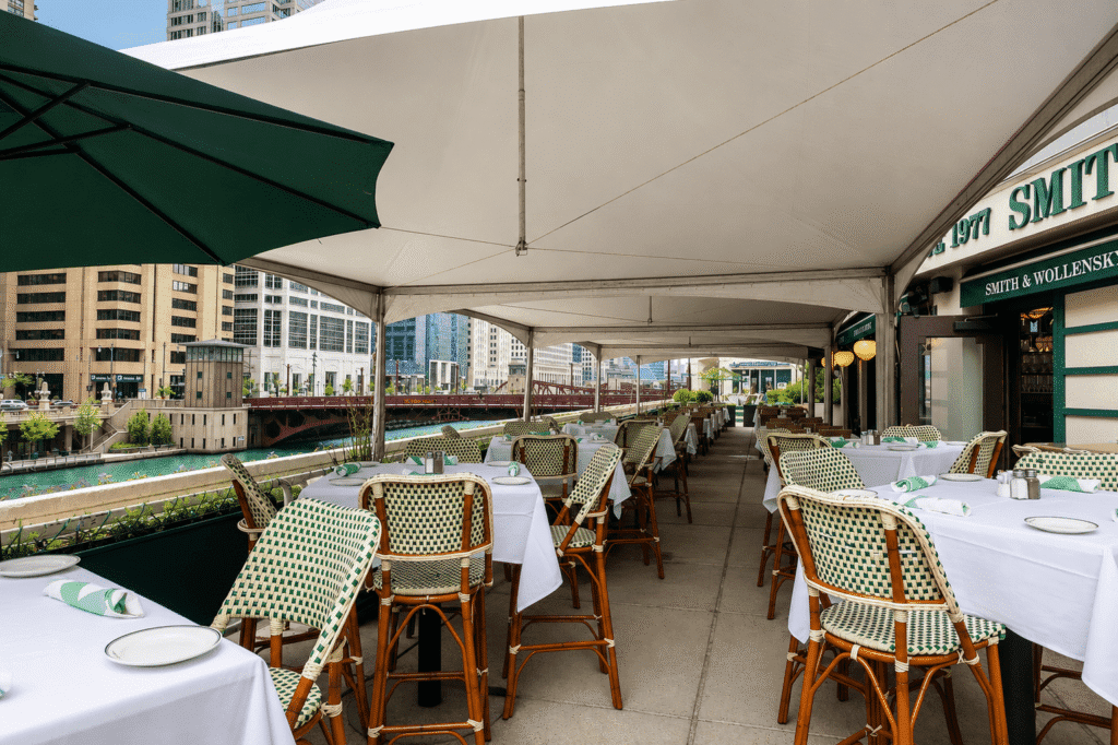 Outdoor restaurant patio with empty tables and chairs under a white canopy, overlooking a river and city buildings, with a green umbrella and place settings on the tables.