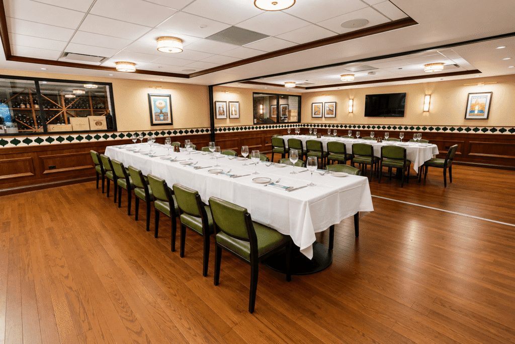 A spacious, elegant dining room with two long tables set with white tablecloths, plates, glasses, and green chairs on a polished wooden floor; walls decorated with framed art and wine storage visible through a window.
