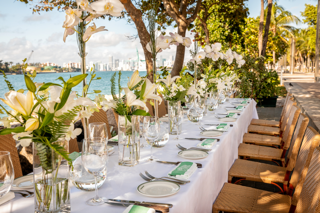 A long outdoor dining table with a white tablecloth is set with plates, glasses, cutlery, and green napkins. Clear vases with white flowers decorate the table, which is beside water and surrounded by trees. City skyline in the background.