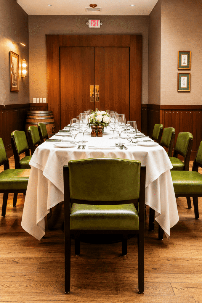 A formal dining table set for eight with green chairs, white tablecloth, glassware, and a flower centerpiece, in a warmly lit room with wooden doors and framed art on the walls.