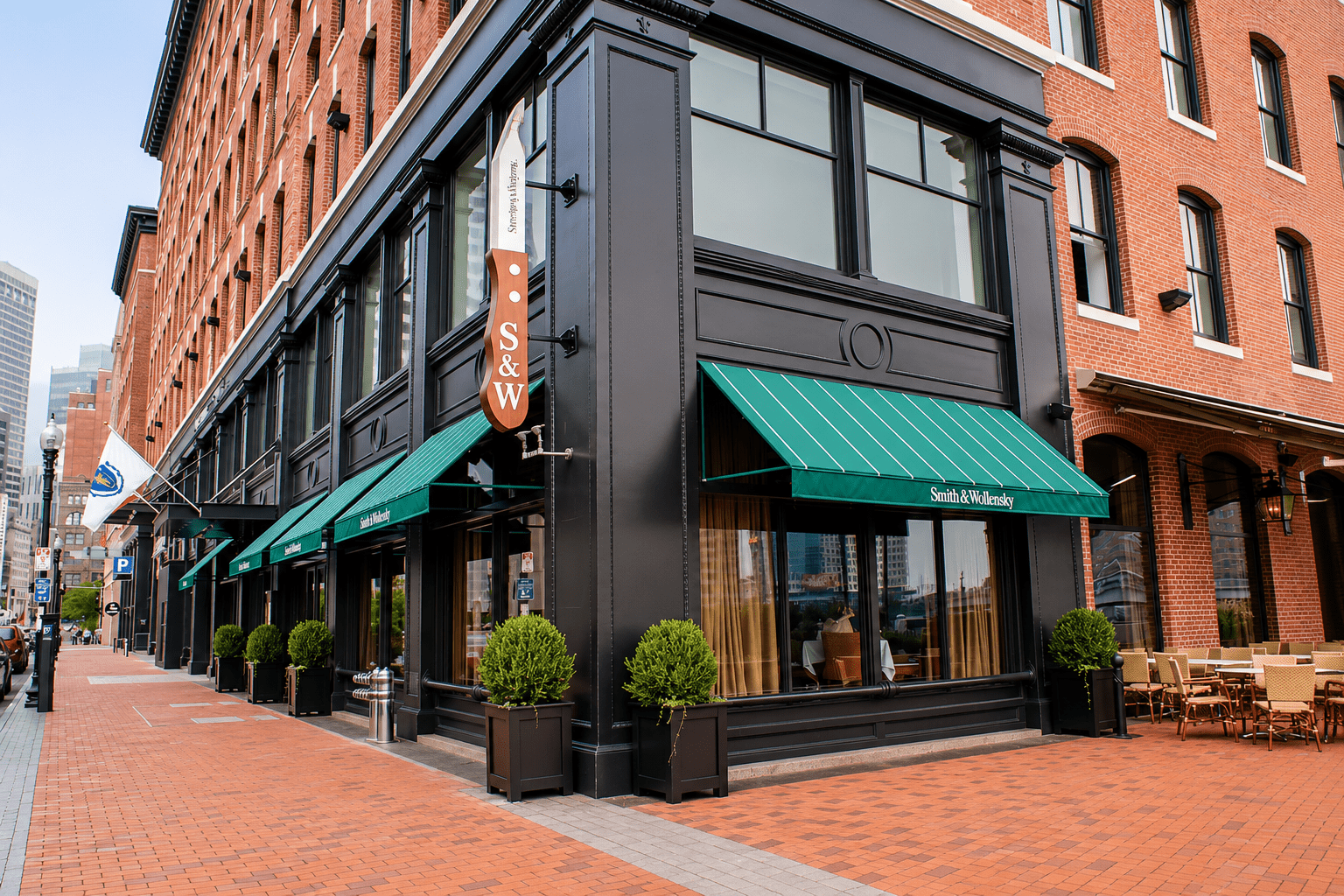 A brick corner building with large windows, green awnings labeled “Smith & Wollensky,” and outdoor seating on a red brick sidewalk in an urban setting.
