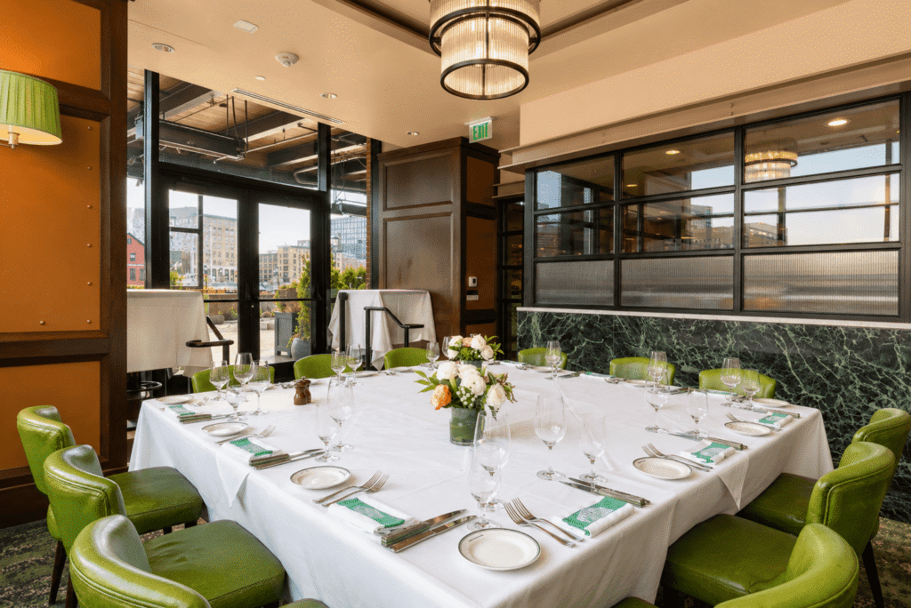A private dining room with a square table set for nine, featuring green leather chairs, white tablecloth, floral centerpiece, and large windows allowing natural light, with city views in the background.