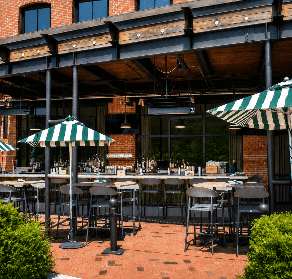 Outdoor bar seating area with high tables and chairs, green-and-white striped umbrellas, and a brick building in the background. The bar is stocked with bottles and glassware, and greenery borders the patio.