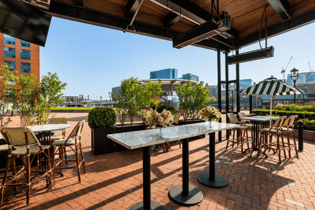 Outdoor patio with marble-topped tables, flower arrangements, wicker chairs, and green plants. Urban buildings and a clear blue sky are visible in the background.