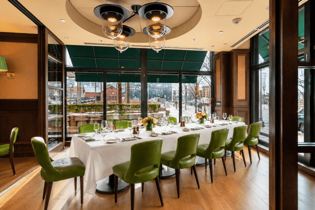 A long dining table with a white tablecloth, set for a meal with green chairs around it. The room has large windows, wood floors, and modern lighting, offering a view of a city street and outdoor patio.