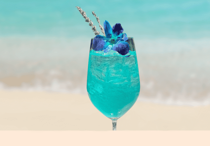 A glass of bright blue cocktail with ice, garnished with a blue flower and lavender sprigs, sits in front of a blurred beach and ocean background.