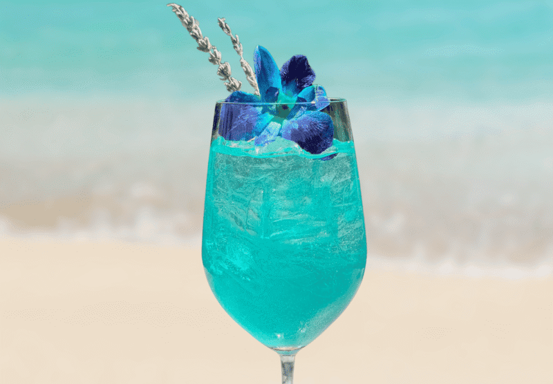 A blue cocktail with ice, garnished with a purple flower and lavender sprig, sits in a glass against a blurred beach and ocean background.