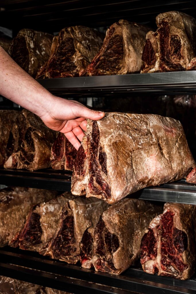 A hand is holding a large piece of dry-aged beef on a metal rack, with several other dry-aged beef cuts stacked on the shelves in the background.